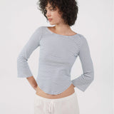 A woman with short curly dark hair wearing a blue and white horizontally striped long-sleeve crop top and white drawstring pants, photographed against a plain light gray studio background.