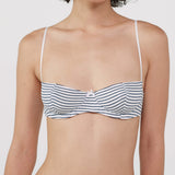 A close-up of a model wearing a white bra with thin shoulder straps and horizontal navy blue stripes, set against a plain light gray background.