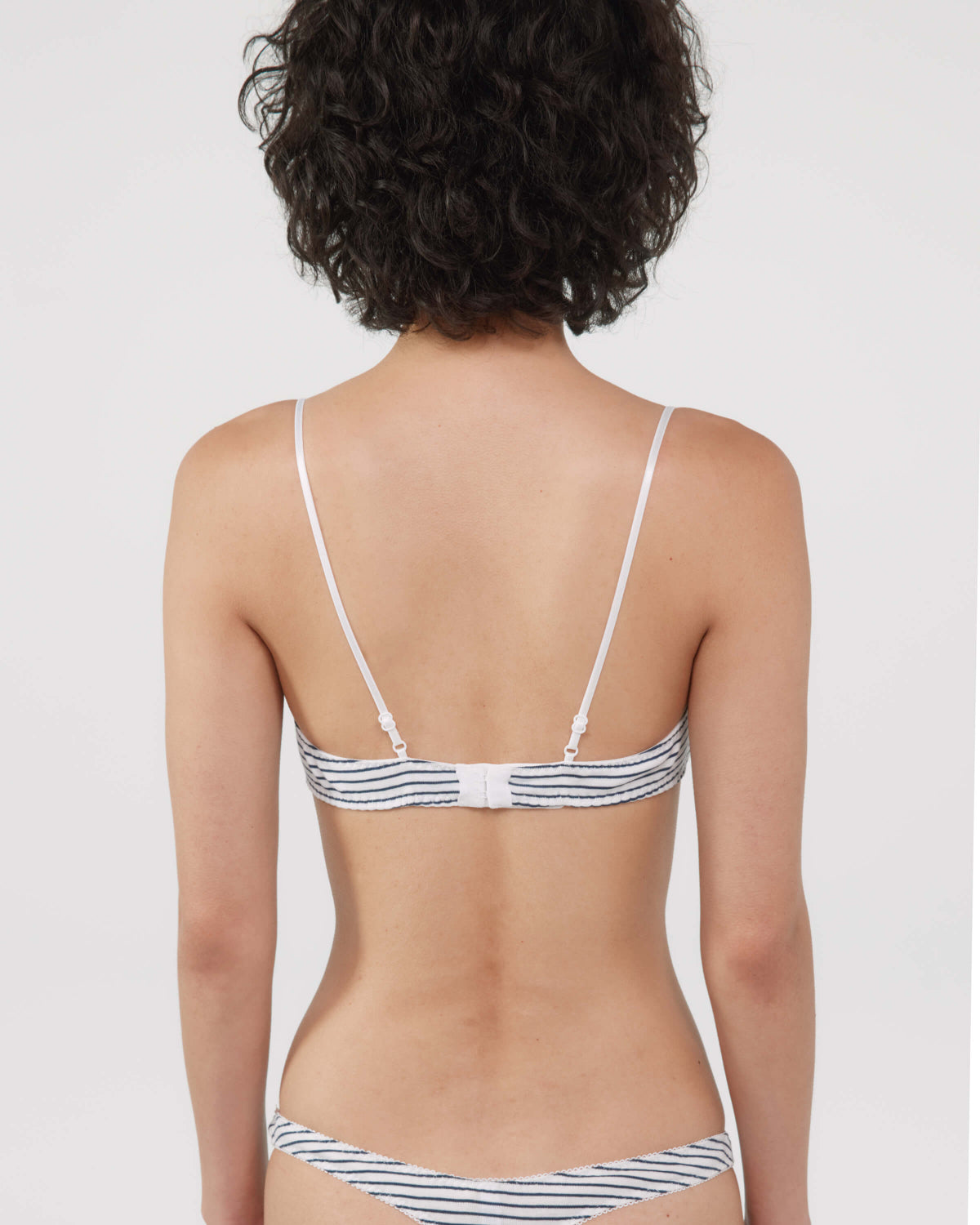 Rear view of a woman wearing a striped bra and thong with thin white straps and navy and white horizontal stripes, against a light gray studio background.